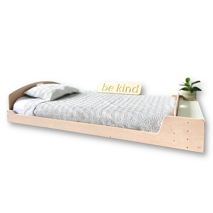 A twin-sized Montessori floor bed made of wood with rounded corners and a 'be kind' sign placed at the top. The bed is accompanied by a pillow and a small potted plant.