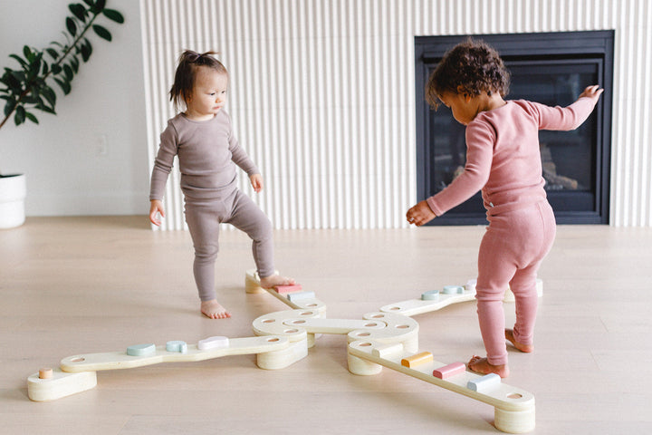 Two children playing with a wooden seesaw in a room with a fireplace and plant.